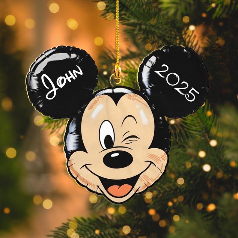 May include: A Mickey Mouse-shaped Christmas ornament with black ears, the left ear displaying the name "John" and the right ear displaying the year "2025". The ornament features a winking Mickey face with a smiling expression. It is hanging from a gold string.