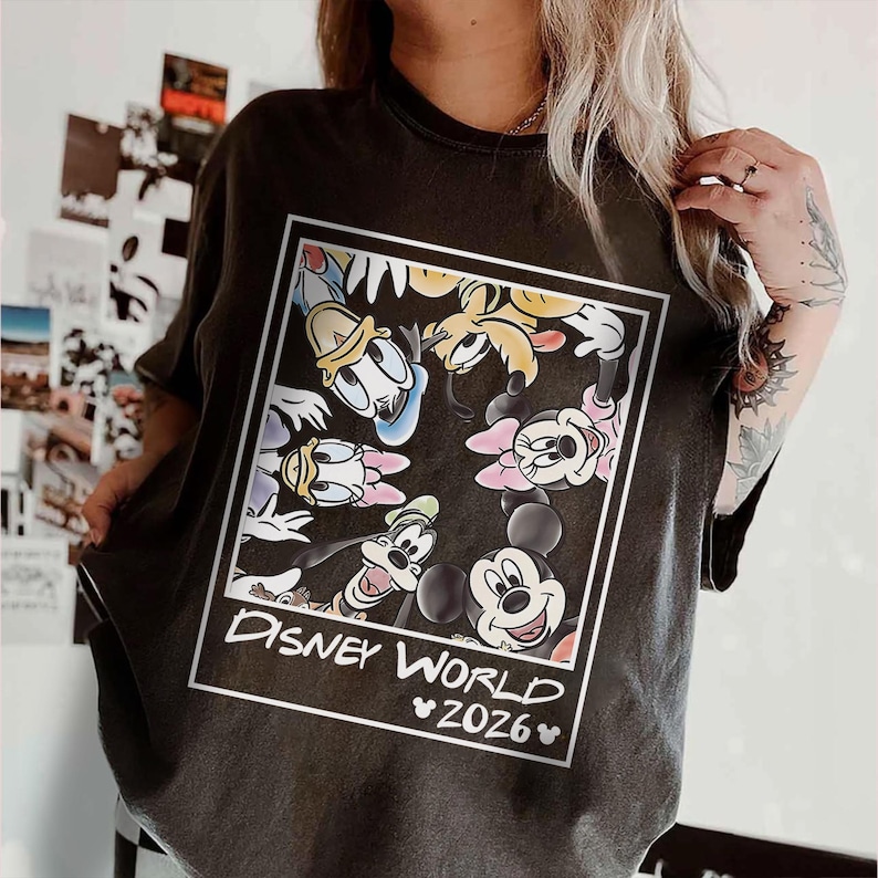 May include: Dark gray t-shirt featuring a framed graphic of Disney characters including Mickey Mouse, Minnie Mouse, Donald Duck, and Goofy. The text "DISNEY WORLD 2026" is printed below the characters in white.