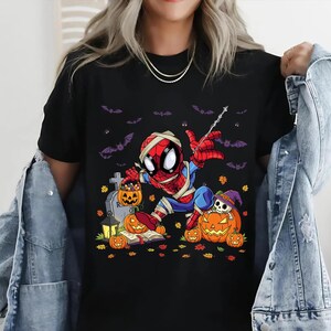 May include: Black t-shirt featuring a Halloween-themed Spider-Man design. The illustration shows Spider-Man in a mummy costume, surrounded by pumpkins, bats, and a tombstone. The graphic is colorful and detailed, perfect for the spooky season.