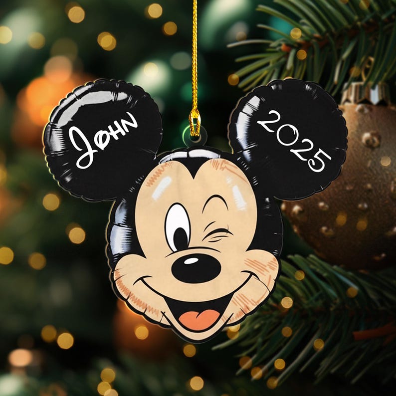 May include: A Mickey Mouse-shaped Christmas ornament with the name "John" on one ear and the year "2025" on the other. The ornament is smiling and winking, with a gold string for hanging. The background includes Christmas tree branches and lights.