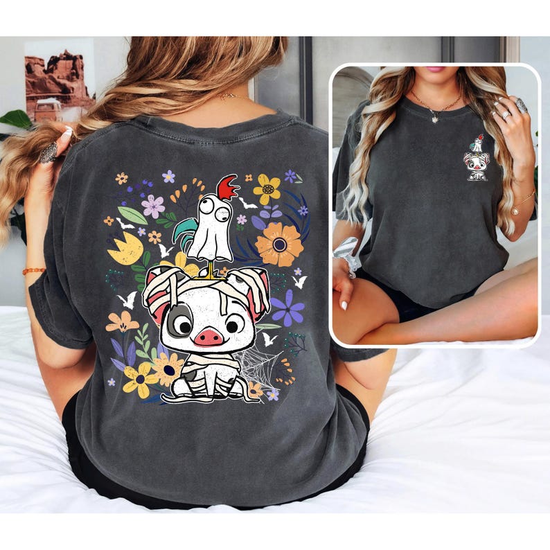 May include: Dark gray t-shirt featuring a cartoon pig and chicken design with colorful flowers. The pig is wrapped in bandages, and the chicken is wearing a ghost costume. The design is on the back of the shirt, with a smaller version on the front.