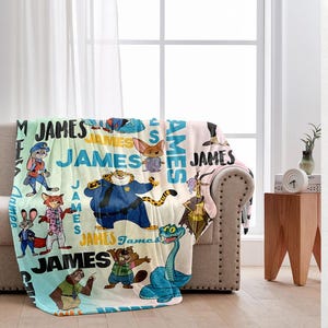K&ouml;nnte beinhalten: Eine personalisierte Fleece-Decke mit Cartoon-Figuren und dem Namen "JAMES" in verschiedenen Schriftarten. Die Decke hat ein hellblaues, gr&uuml;nes und rosa Farbschema mit Bildern von Tieren in verschiedenen Outfits.