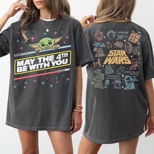 K&ouml;nnte beinhalten: Dunkelgraues T-Shirt mit einem Baby Yoda-Motiv, Lichtschwertern und dem Text "MAY THE 4TH BE WITH YOU". Auf der R&uuml;ckseite sind Star Wars-Charaktere und Zitate abgebildet. L&auml;ssiger, &uuml;bergro&szlig;er Schnitt.