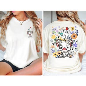 May include: Cream-colored t-shirt featuring a cartoon pig character with a ghost and a rooster. The front of the shirt has a small graphic, while the back showcases a larger design with flowers and bats. The shirt is a casual, comfortable style.