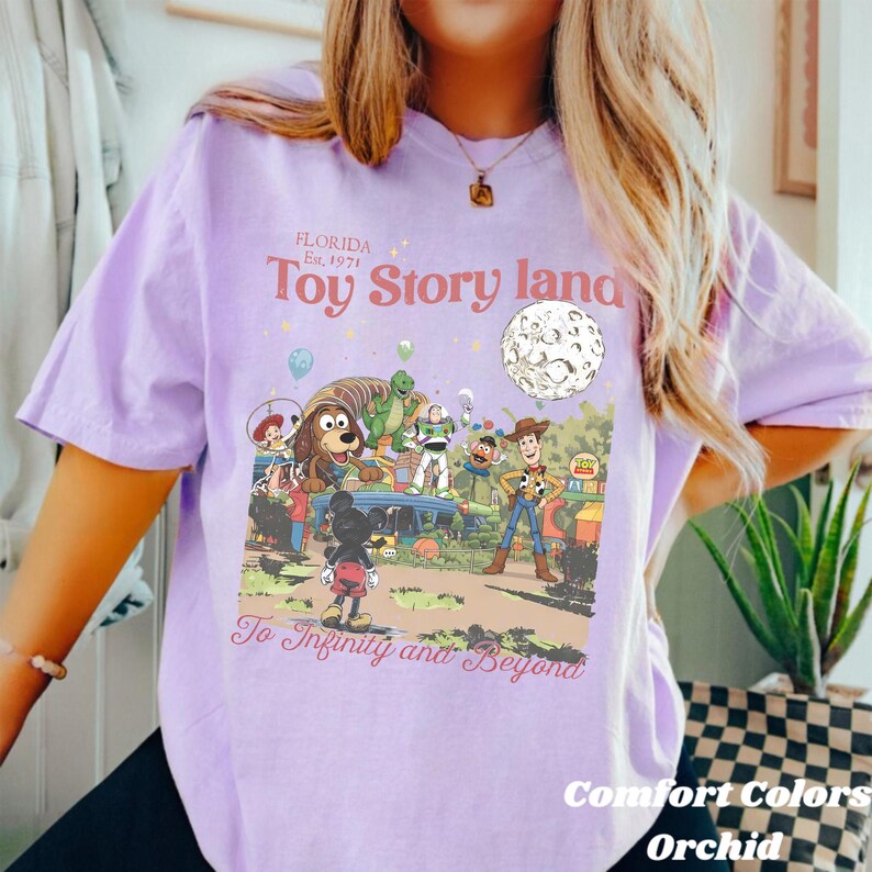 May include: A Comfort Colors orchid-colored t-shirt featuring a graphic of Toy Story characters and the text "Toy Story Land" and "To Infinity and Beyond". The design includes Woody, Buzz Lightyear, and other characters. The shirt is a light purple color.