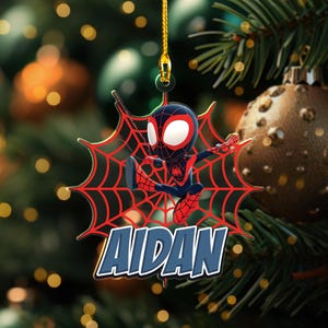 May include: A Christmas tree ornament featuring a cartoon Spider-Man character in a dynamic pose, set against a red spiderweb design. The name "AIDAN" is displayed in blue and white. The ornament hangs from a gold string.