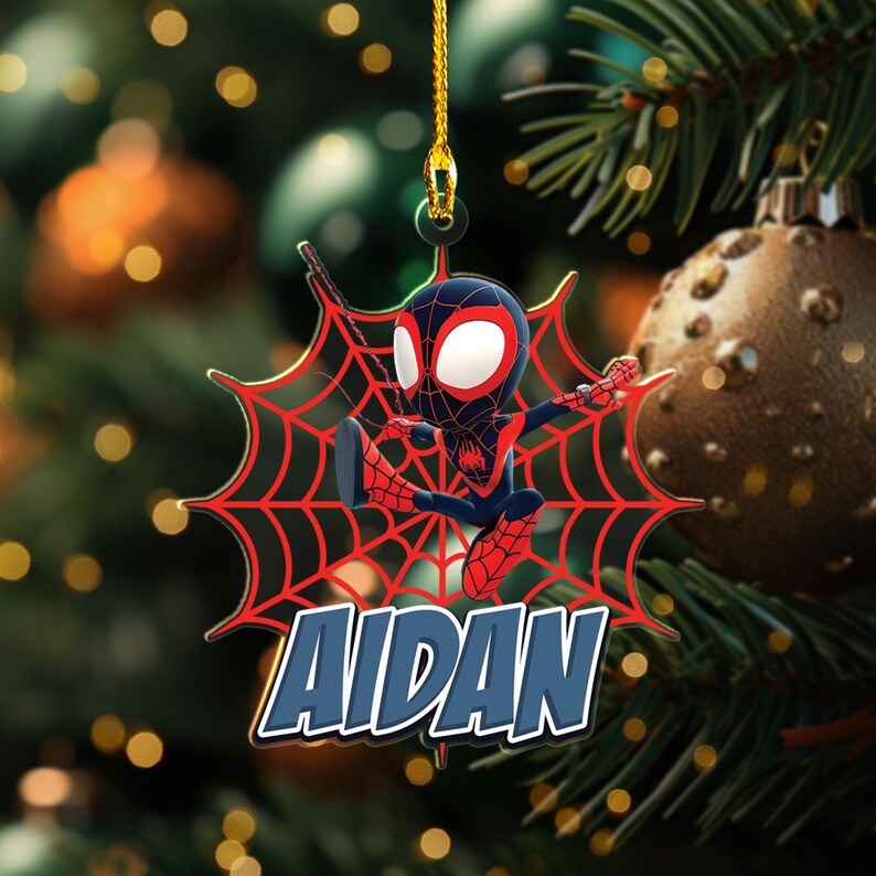 May include: A Christmas tree ornament featuring a cartoon Spider-Man character in a dynamic pose, set against a red spiderweb design. The name "AIDAN" is displayed in blue and white. The ornament hangs from a gold string.