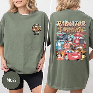 Puede incluir: Camiseta verde musgo con un dise&ntilde;o de coche de dibujos animados en la parte delantera y trasera. La parte delantera tiene un solo coche en un c&iacute;rculo a cuadros con la palabra "NAME" debajo. La parte trasera muestra el texto "RADIATOR SPRINGS" e ilustraciones de varios coches.