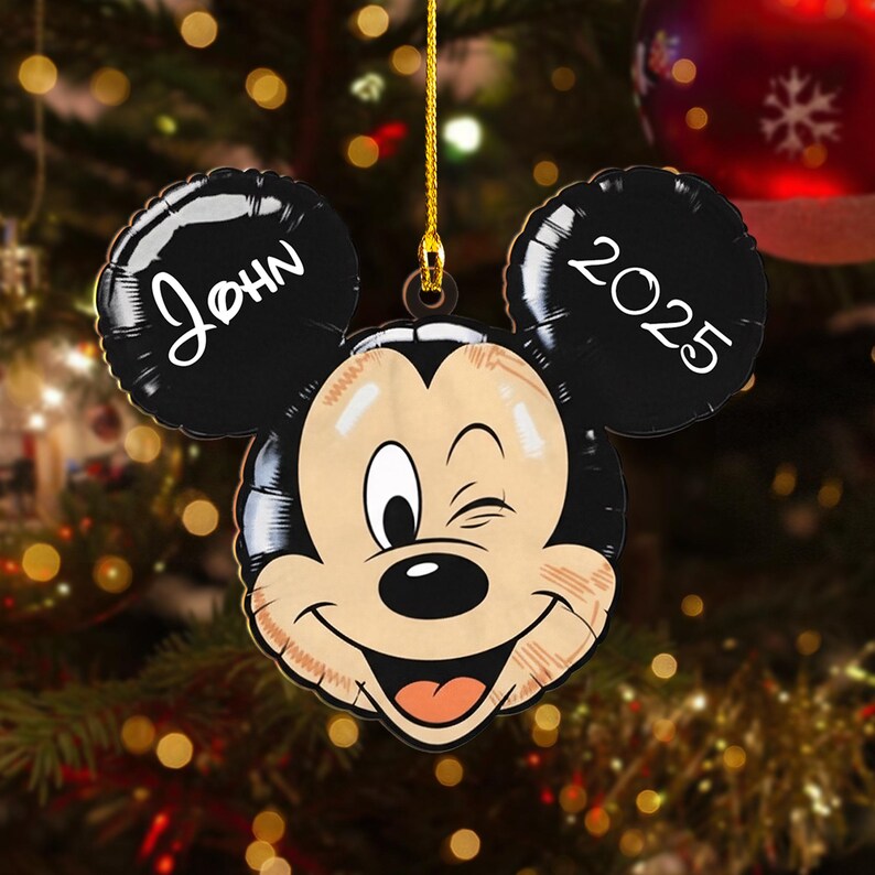 May include: A Mickey Mouse-shaped Christmas ornament with the name "John" on one ear and the year "2025" on the other. The ornament is smiling and winking, with a gold string for hanging. The background is a blurred Christmas tree.