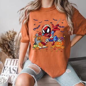 May include: Orange short-sleeved t-shirt featuring a Halloween-themed Spider-Man graphic. The design includes a mummy-wrapped Spider-Man, pumpkins, bats, and a tombstone. The shirt is a comfortable, casual style.