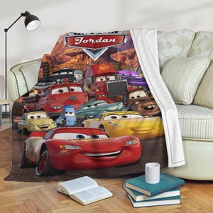May include: A fleece blanket featuring a colorful design of various Cars characters. The blanket has a red, white, and blue color scheme, with the name "Jordan" displayed at the top. The blanket is draped over a sofa.