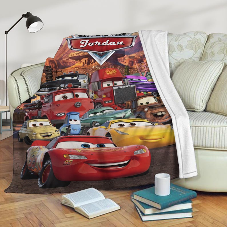 May include: A fleece blanket featuring a colorful design of various Cars characters. The blanket has a red, white, and blue color scheme, with the name "Jordan" displayed at the top. The blanket is draped over a sofa.
