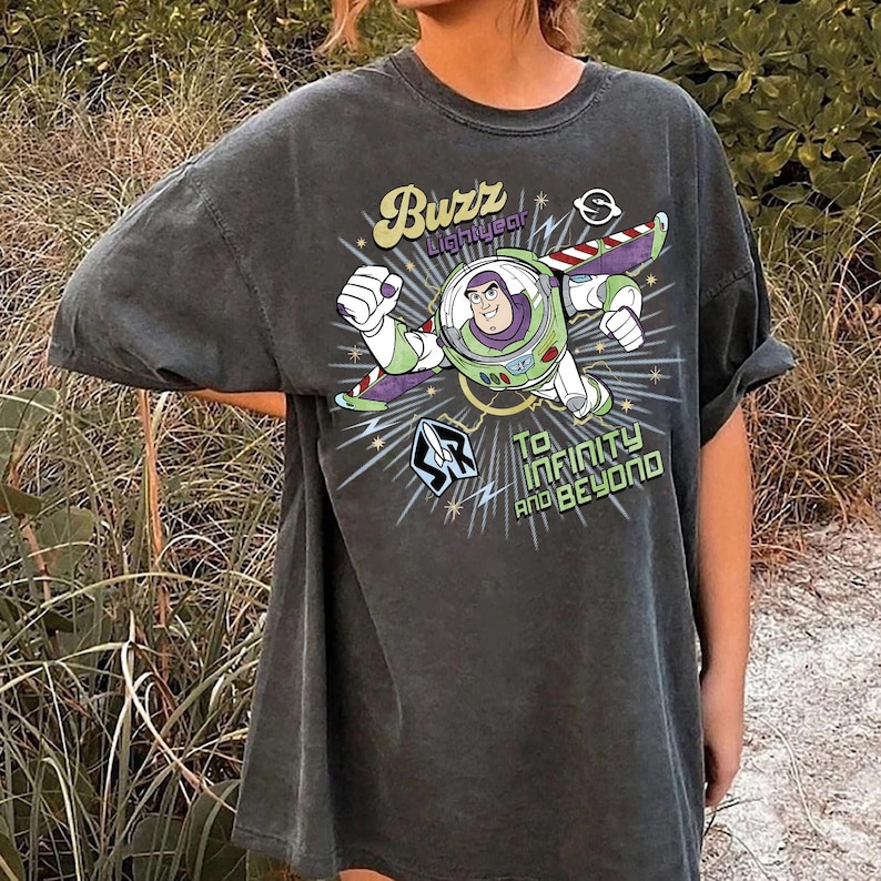 Puede incluir: Camiseta gris oscuro con un dise&ntilde;o de Buzz Lightyear volando. El dise&ntilde;o incluye el texto "Buzz Lightyear" y "To Infinity and Beyond" en una fuente estilizada. La camiseta tiene mangas cortas y un corte holgado.