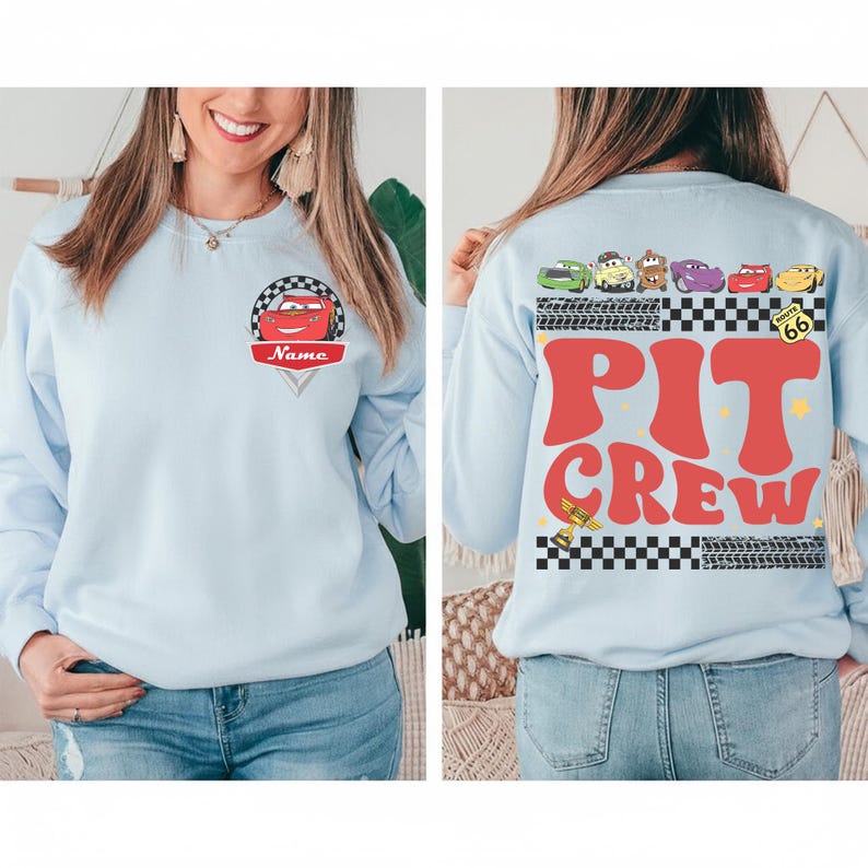 May include: Light blue sweatshirt featuring a cartoon car design with the name "Name" on the front. The back of the sweatshirt displays the words "PIT CREW" in red, along with car graphics and a checkered flag design. A Route 66 sign is also visible.