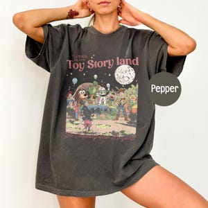 May include: Pepper-colored t-shirt featuring a colorful graphic of Toy Story characters with the text "Toy Story land" and "To Infinity and Beyond". The shirt has a vintage, distressed look.
