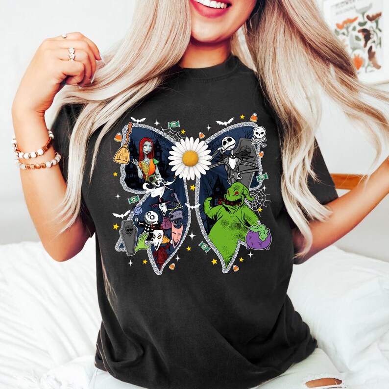 May include: Black t-shirt with a bow design featuring characters from The Nightmare Before Christmas. The bow has a daisy in the centre and characters like Sally, Jack Skellington, and Oogie Boogie. The design includes bats, stars, and candy corn.