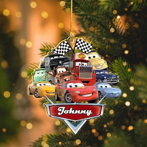 May include: A Cars-themed Christmas ornament featuring Lightning McQueen and other characters. The ornament has a gold string for hanging and a nameplate that says "Johnny". The ornament includes checkered flags.