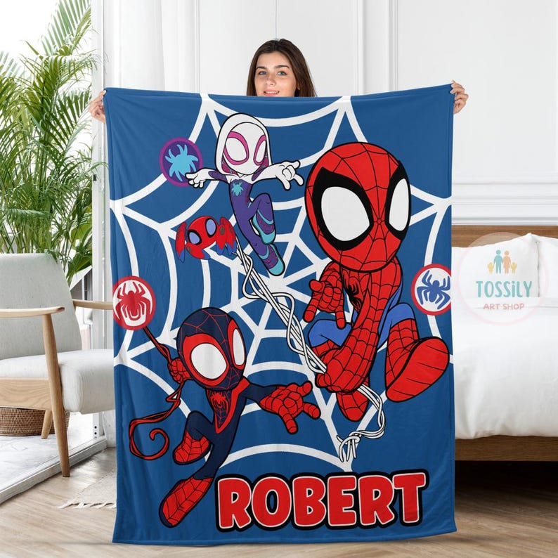 K&ouml;nnte beinhalten: Eine blaue Fleece-Decke mit einem Spinnennetz-Design, das Spider-Man, Spider-Gwen und Miles Morales zeigt. Die Decke tr&auml;gt den Namen "ROBERT", der unten in Rot gedruckt ist.