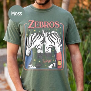 May include: Moss green t-shirt with a graphic of two zebras and the word "ZEBROS" in pink. The zebras are in black and white, with blue and red accents. The shirt has a crew neck.