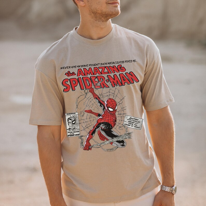 Pu&ograve; includere: T-shirt beige con grafica di Spider-Man in azione, con la scritta "The Amazing Spider-Man". Sono presenti anche vignette di fumetti e la frase "Never has anyone fought such merciless foes as..."