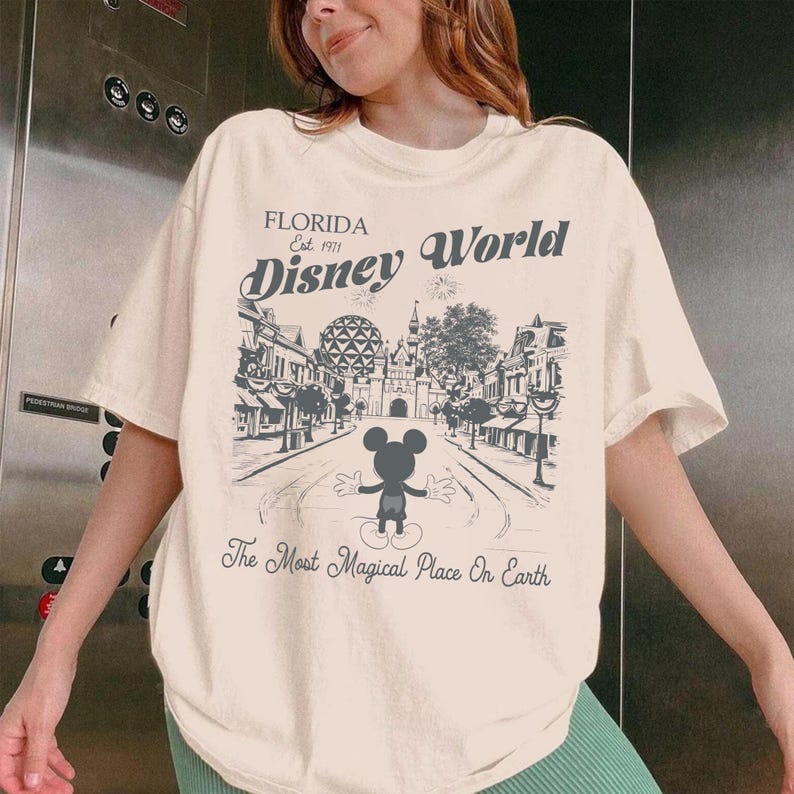 May include: Cream-coloured t-shirt with a black and white illustration of Disney World in Florida. The design includes Mickey Mouse, the text "Florida Est. 1971 Disney World," and the phrase "The Most Magical Place On Earth."