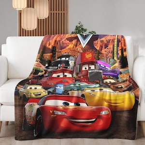 May include: A plush blanket featuring a colorful graphic of characters from the animated movie "Cars." The blanket showcases Lightning McQueen and other vehicles against a desert landscape background. The blanket is draped over a white sofa.