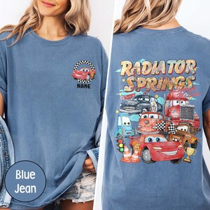 Puede incluir: Camiseta azul vaquera con un dise&ntilde;o de coche de dibujos animados en la parte delantera y un gr&aacute;fico de "Radiator Springs" en la parte trasera. El dise&ntilde;o incluye coches de dibujos animados, banderas a cuadros y el texto "NAME".