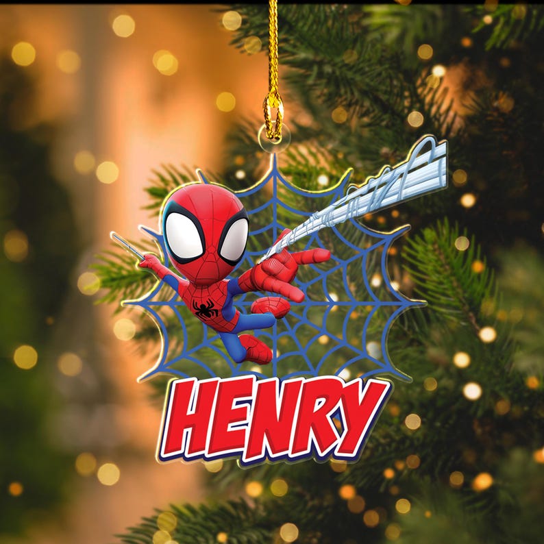 May include: A red, blue, and black Spider-Man ornament with the name "HENRY" in red letters. The ornament is hanging from a gold string and features a blue spider web design. The background is a blurred image of a Christmas tree.