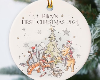 Puede incluir: Adorno navideño redondo y blanco con una cuerda dorada. Presenta una ilustración de dibujos animados de Winnie the Pooh y sus amigos decorando un árbol de Navidad. El texto dice "Riley's FIRST CHRISTMAS 2024."