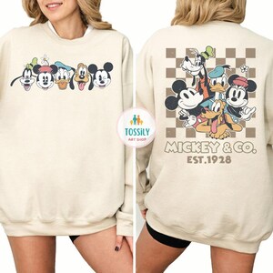 May include: Cream-colored sweatshirt featuring a cartoon graphic of Mickey Mouse, Goofy, Donald Duck, and Pluto. The front displays the characters' faces, while the back showcases a group image with a checkered background and the text "Mickey & Co. EST.1928."