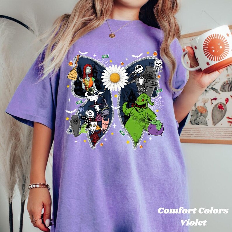 May include: Violet Comfort Colors t-shirt with a butterfly design featuring characters from The Nightmare Before Christmas. The design includes Sally, Jack Skellington, Oogie Boogie, and other characters. A large white daisy is in the center.