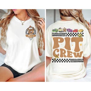 May include: Cream-colored t-shirt featuring a cartoon car design with the text "Name" on the front. The back of the shirt displays the words "PIT CREW" in brown, along with car graphics and a Route 66 sign.