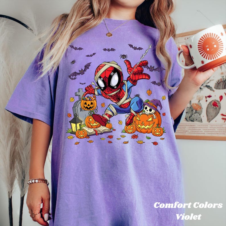 May include: Violet Comfort Colors t-shirt featuring a Halloween-themed Spider-Man graphic. The design includes pumpkins, bats, and a tombstone. The text "Comfort Colors Violet" is at the bottom of the shirt.