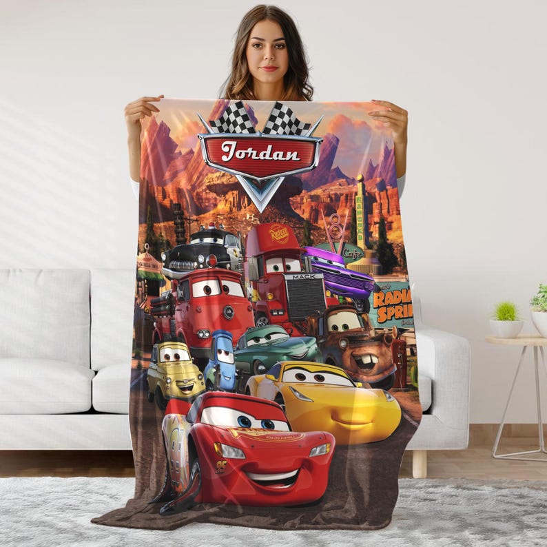 May include: A fleece blanket featuring characters from the animated movie "Cars." The blanket has a colorful design with Lightning McQueen and other characters. The name "Jordan" is displayed on the blanket.