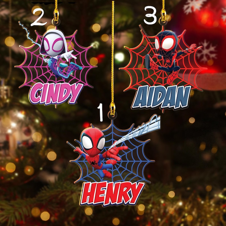 May include: Three superhero-themed Christmas ornaments. Each ornament features a character on a spiderweb background with a name. The names are "Henry", "Cindy", and "Aidan". The ornaments are red, blue, and pink.