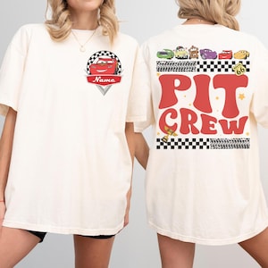 May include: Cream-colored t-shirt featuring a red race car graphic with the name "Name" and a checkered flag design. The back of the shirt displays "PIT CREW" in red, along with various cartoon race cars and Route 66 signage.