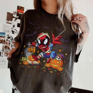 May include: Dark gray t-shirt featuring a Halloween-themed Spider-Man graphic. The cartoon Spider-Man is dressed as a mummy and surrounded by pumpkins, bats, and a tombstone. The design is colorful and whimsical, perfect for the spooky season.