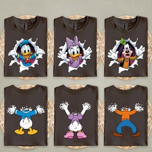 May include: Six dark brown t-shirts with cartoon character graphics. Donald Duck, Daisy Duck, and Goofy are featured, each appearing to burst through a hole. The shirts are arranged on a woven surface.