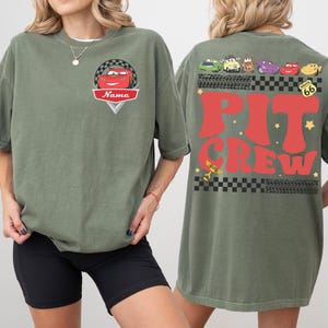 May include: Olive green t-shirt featuring a cartoon car design with the word "Name" on the front. The back of the shirt displays the words "Pit Crew" in red, along with various car graphics and Route 66 signage.