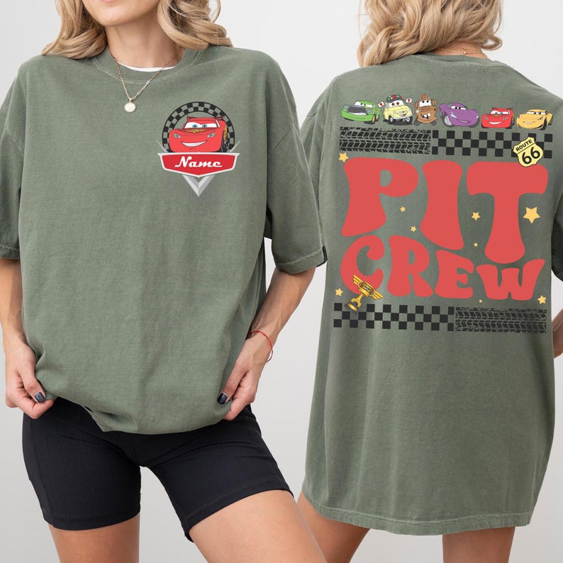 May include: Olive green t-shirt featuring a cartoon car design with the word "Name" on the front. The back of the shirt displays the words "Pit Crew" in red, along with various car graphics and Route 66 signage.