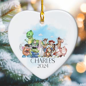 May include: White heart-shaped ceramic ornament featuring a watercolour illustration of the Toy Story characters. The characters include Woody, Buzz Lightyear, Jessie, Rex, Slinky Dog, Mr. and Mrs. Potato Head, and Hamm. The ornament is personalised with the name "CHARLES" and the year "2024".