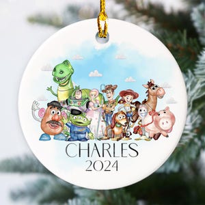 May include: A white ceramic Christmas bauble featuring a watercolour illustration of the Toy Story characters, including Woody, Buzz Lightyear, Rex, and Mr. Potato Head. The bauble is personalised with the name "CHARLES" and the year "2024".