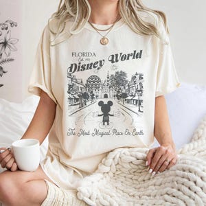 May include: Cream-coloured t-shirt with a black and white graphic of Disney World in Florida. The design includes Mickey Mouse, the park's name, and the phrase "The Most Magical Place On Earth."