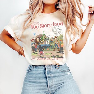 May include: Cream-colored t-shirt featuring a vintage-style graphic of Toy Story characters in a theme park setting. The text "Toy Story land" and "To Infinity and Beyond" are also visible. The shirt is paired with blue jeans.