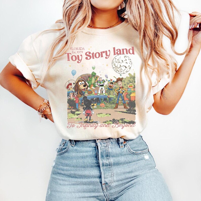 May include: Cream-colored t-shirt featuring a vintage-style graphic of Toy Story characters in a theme park setting. The text "Toy Story land" and "To Infinity and Beyond" are also visible. The shirt is paired with blue jeans.