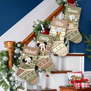 May include: Five Christmas stockings with personalized names and Disney character designs, including Mickey Mouse and Donald Duck. The stockings are in shades of brown and green with a festive pattern. They are displayed on a staircase decorated with garland.