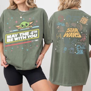 K&ouml;nnte beinhalten: Olivgr&uuml;nes T-Shirt mit einer Grafik von Grogu, Lichtschwertern und dem Text "MAY THE 4TH BE WITH YOU". Auf der R&uuml;ckseite des Shirts befinden sich Star Wars-Grafiken und Text, darunter "STAR WARS" und "THIS IS THE WAY".