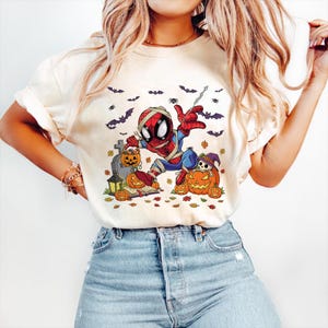 May include: Cream-colored t-shirt featuring a Halloween-themed Spider-Man graphic. The design includes a mummy-wrapped Spider-Man, pumpkins, bats, and a tombstone. The shirt is paired with blue jeans.