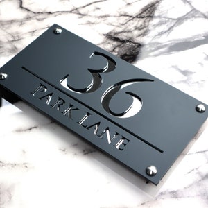 Puede incluir: Un letrero de número de casa de acrílico negro con el número 36 y el nombre de la calle "Park Lane" recortados. El letrero tiene un diseño moderno y minimalista.