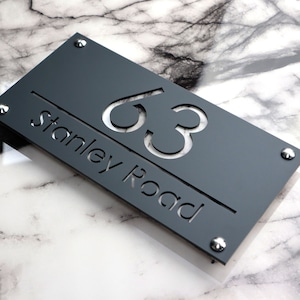 May include: A dark grey rectangular house sign with the number '63' cut out, and the words 'Stanley Road'. The sign is fixed with four silver screws. The background features a white and grey marble effect.